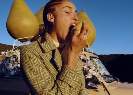Adwoa Aboah met gevlochten haar, bijtend in een peer met op de achtergrond drie grote peren op een autorek.